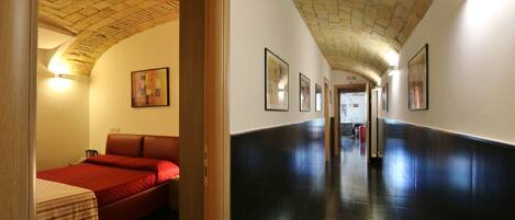 A hotel corridor with a tiled floor, a bed with a red bedspread, and framed artwork on the walls.