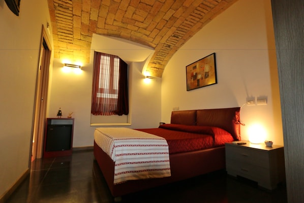 A bedroom with a brick ceiling, a bed with red bedding, a bedside table with a lamp, and a small cabinet. - Residenza Adriana (Rome)