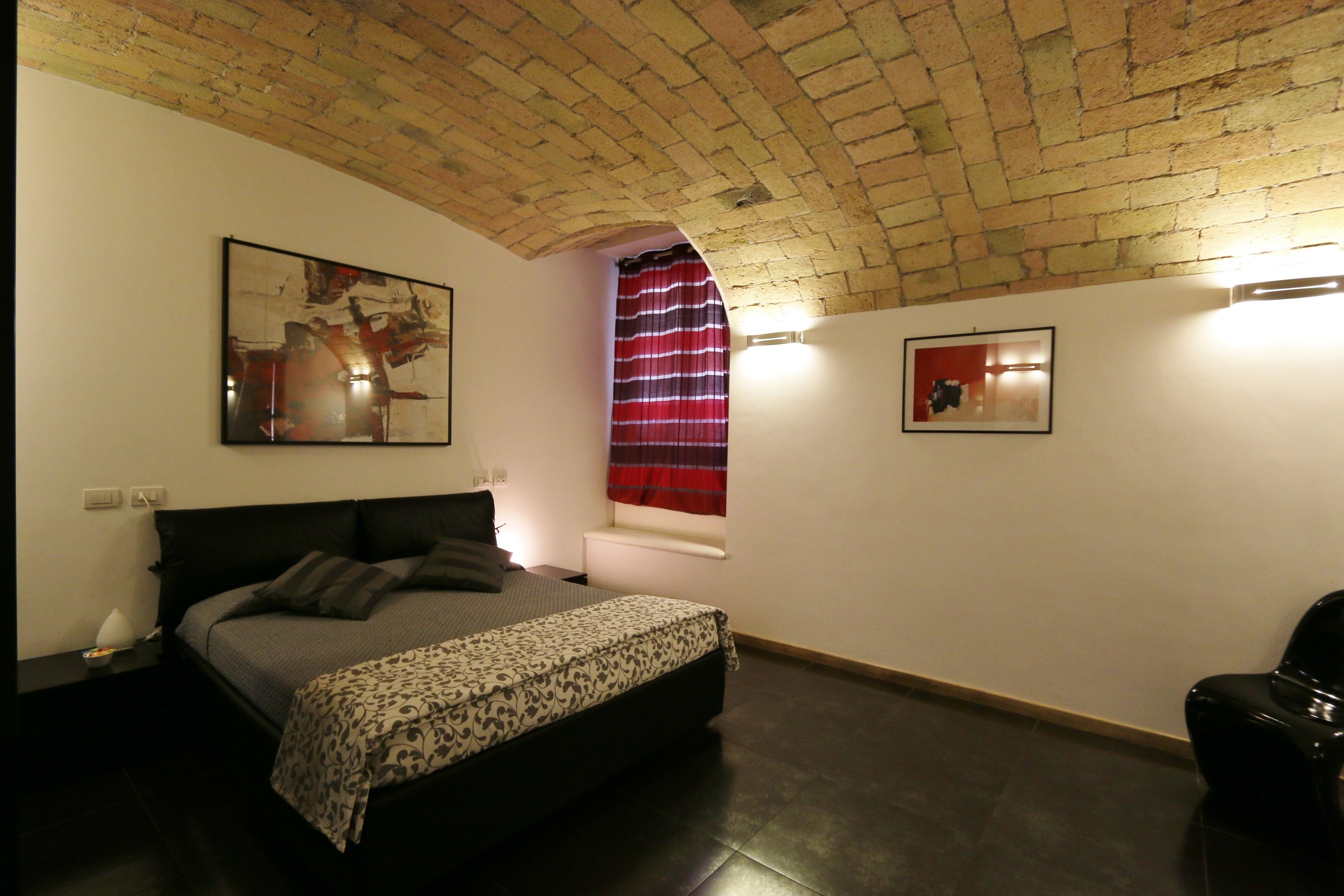 A bedroom with a brick ceiling, a bed with a patterned bedspread, a window with a red curtain, and two framed pictures on the walls.