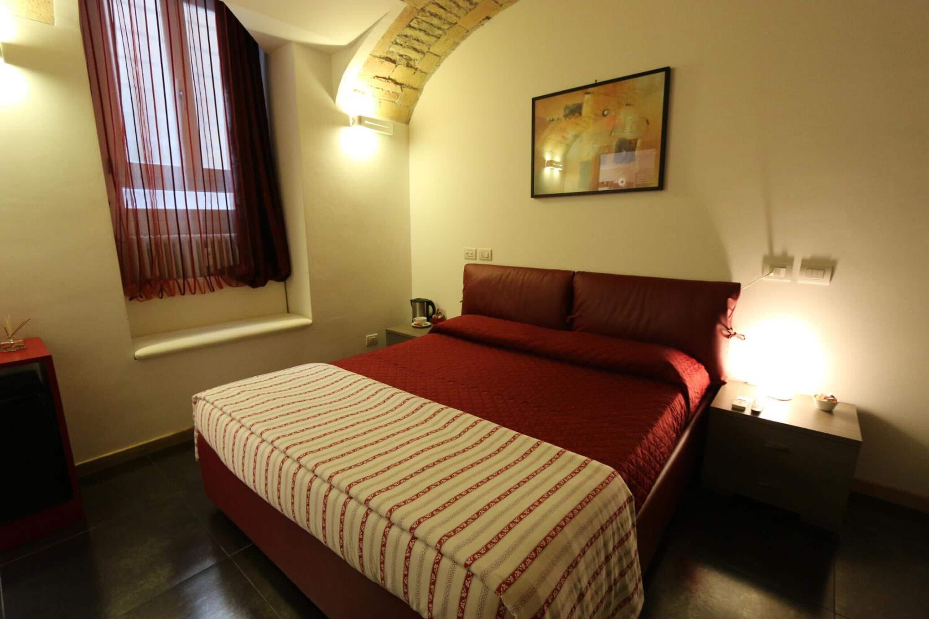A bedroom with a bed, a nightstand, a lamp, a window with curtains, and a painting on the wall.