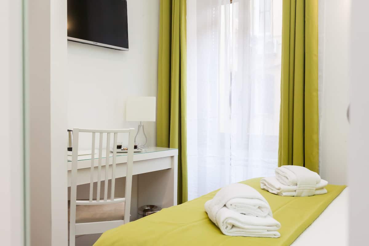 Une chambre d’hôtel comprenant un lit, un bureau avec une chaise, un téléviseur fixé au mur et une fenêtre avec des rideaux.