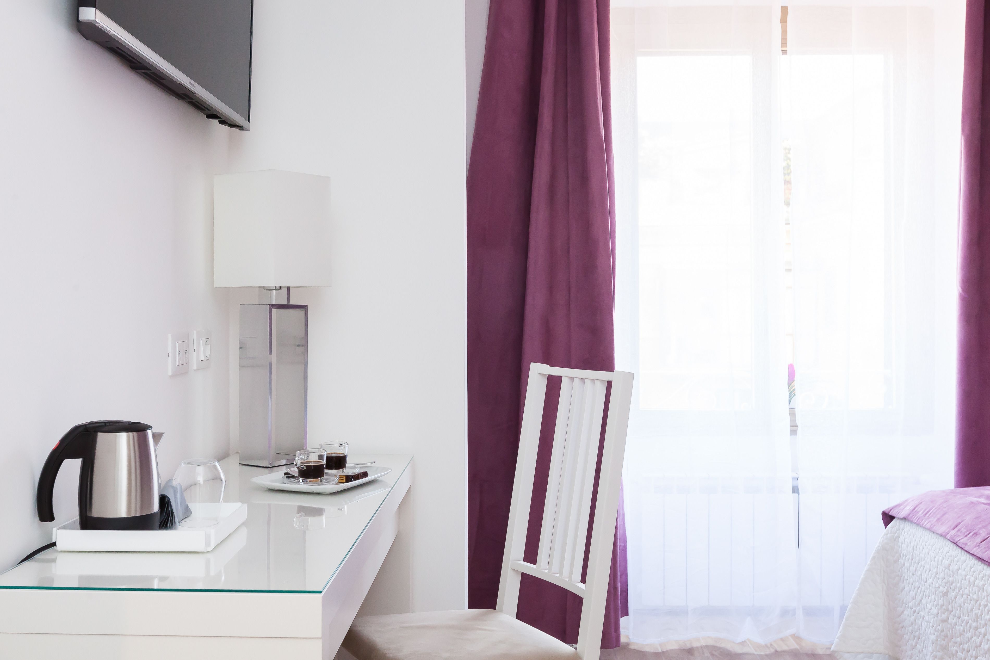 A hotel room with a glass table, a white chair, a bed with a white and purple bedspread, a TV mounted on the wall, and a window with purple curtains.