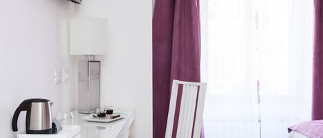 A hotel room with a glass table, a white chair, a bed with a white and purple bedspread, a TV mounted on the wall, and a window with purple curtains.