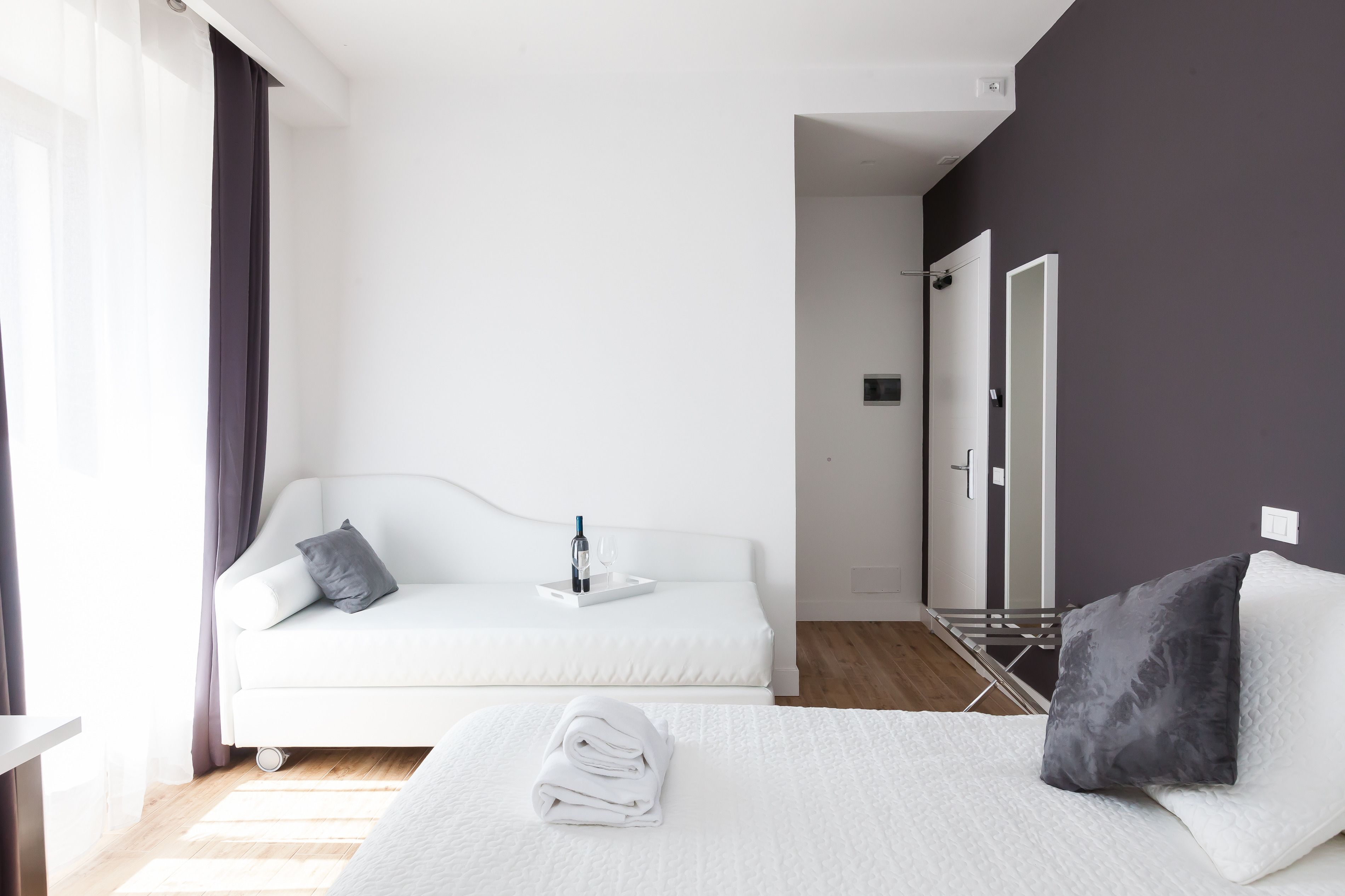 A hotel room with a white bed, a white sofa, a small table with a bottle and glasses, and a dark wall.
