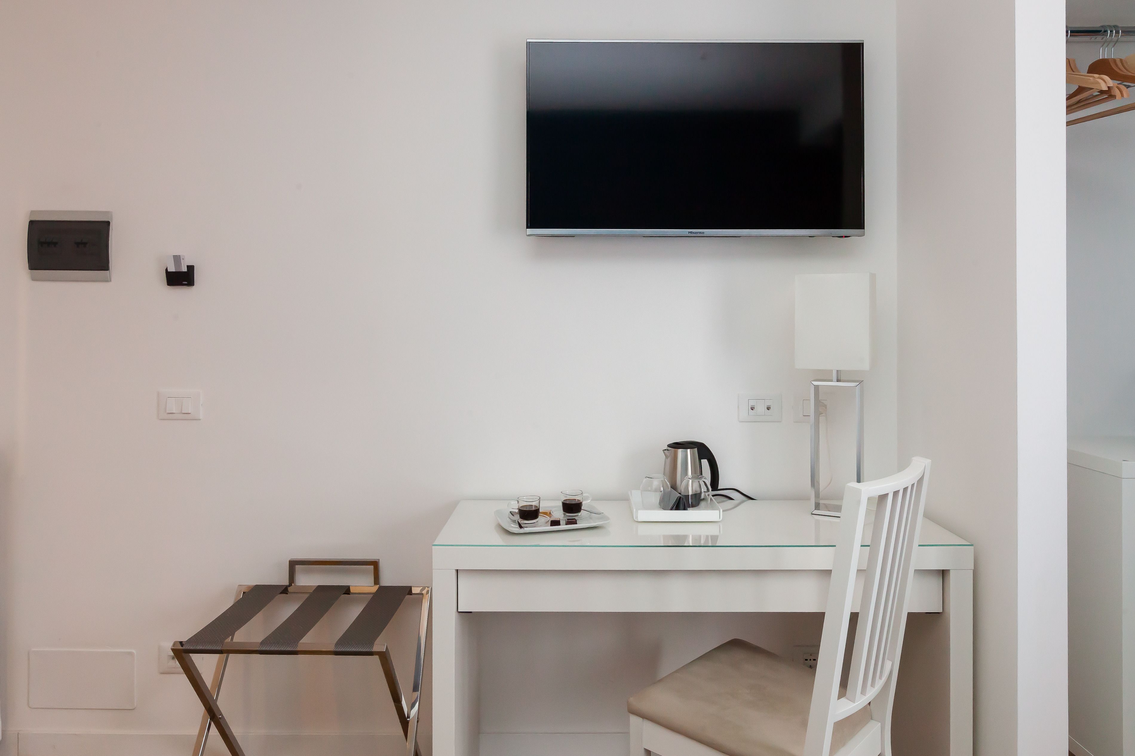 A hotel room with a flat-screen TV mounted on the wall, a small table with a tray of drinks and a coffee pot, a chair, and a coat rack with hanging clothes.