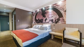 A modern hotel room with a large bed, a nightstand, a chair, and a wall mural of a bridge.