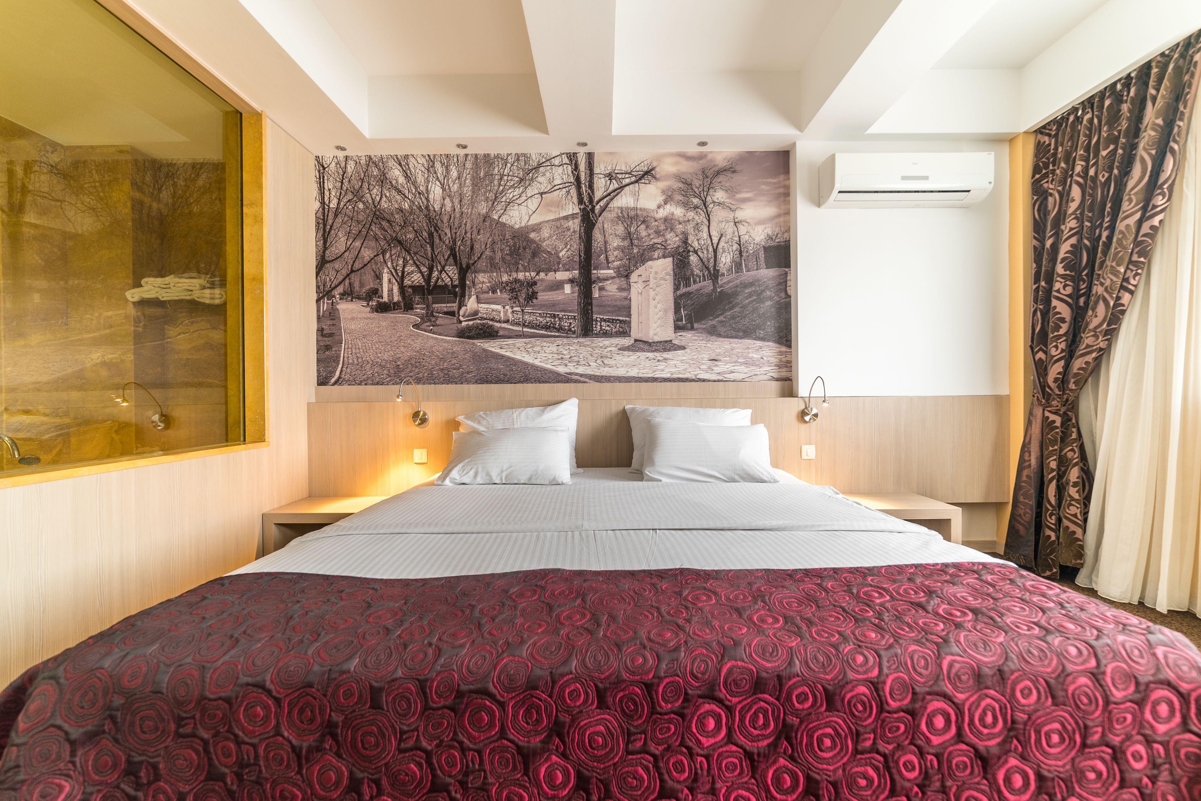A hotel room with a large bed, a nightstand, a wall-mounted air conditioner, and a large window with a view of trees and a building.