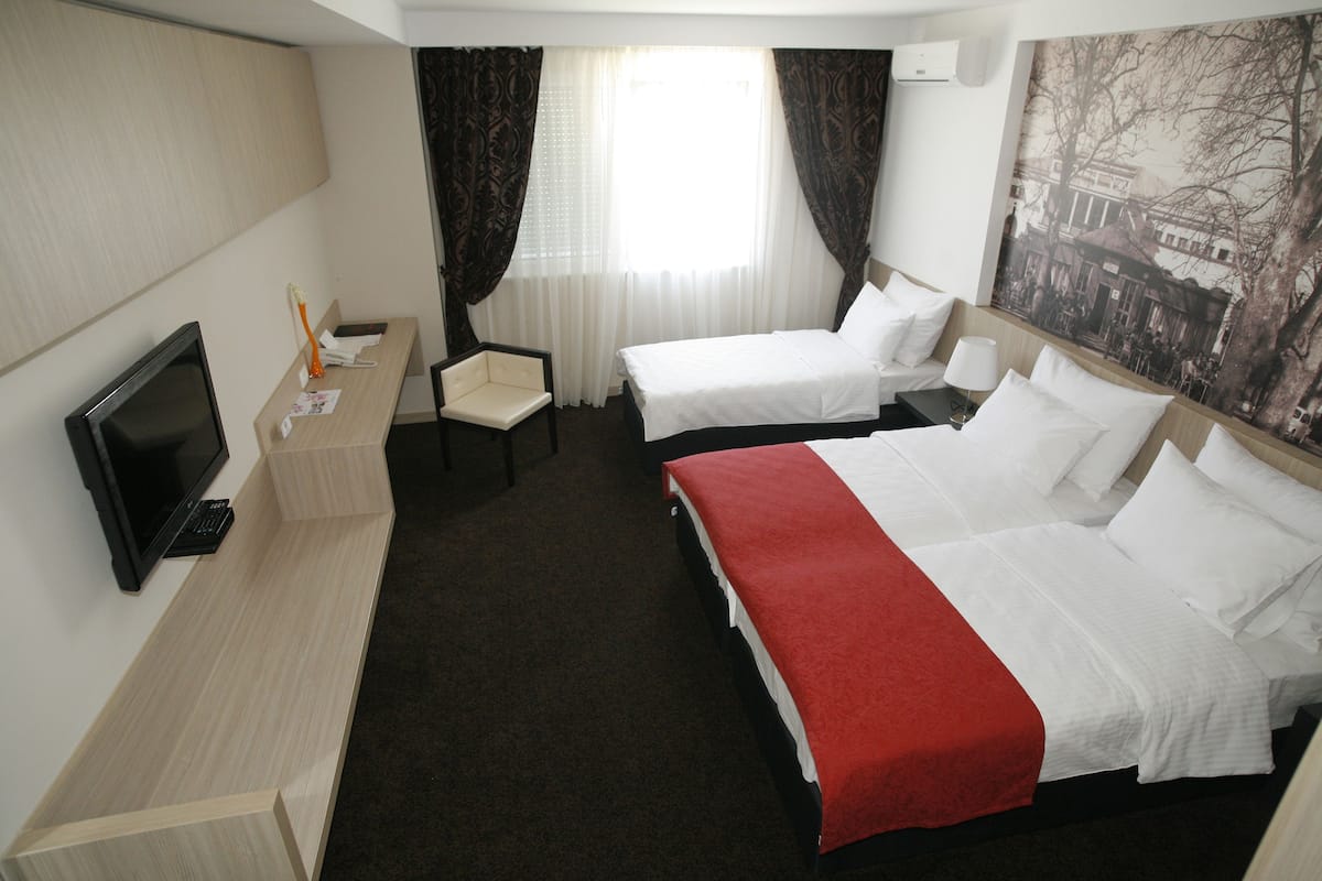Ein Hotelzimmer mit zwei Betten, einem Schreibtisch mit Computer, einem Fernseher und einem großen Fenster mit Vorhängen.