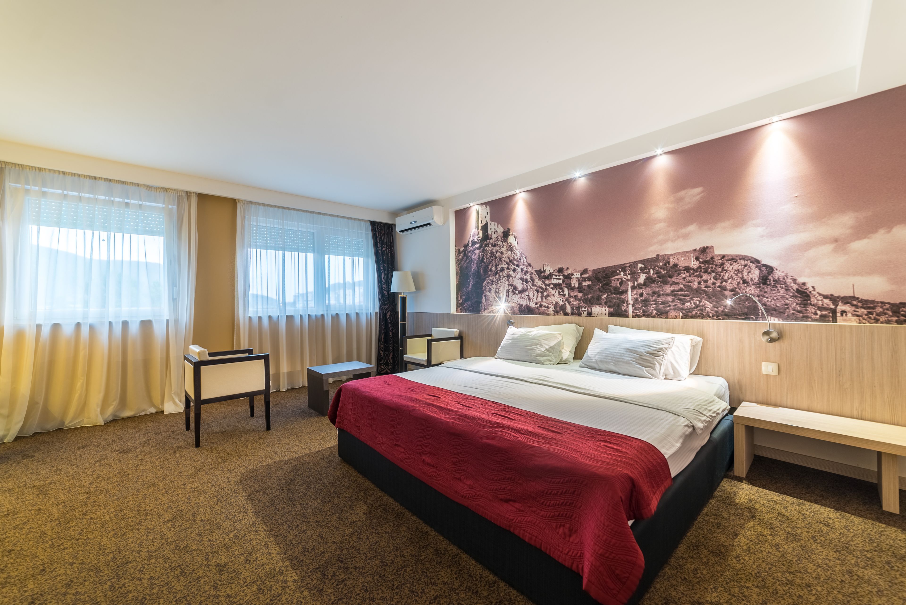 A hotel room with a large bed, a desk, a chair, a lamp, and a wall mural.