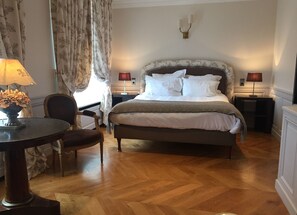 Een netjes ingerichte hotelkamer met een groot bed, twee bedlampen, een houten stoel, een klein rond tafeltje met een lamp en een decoratieve vaas.