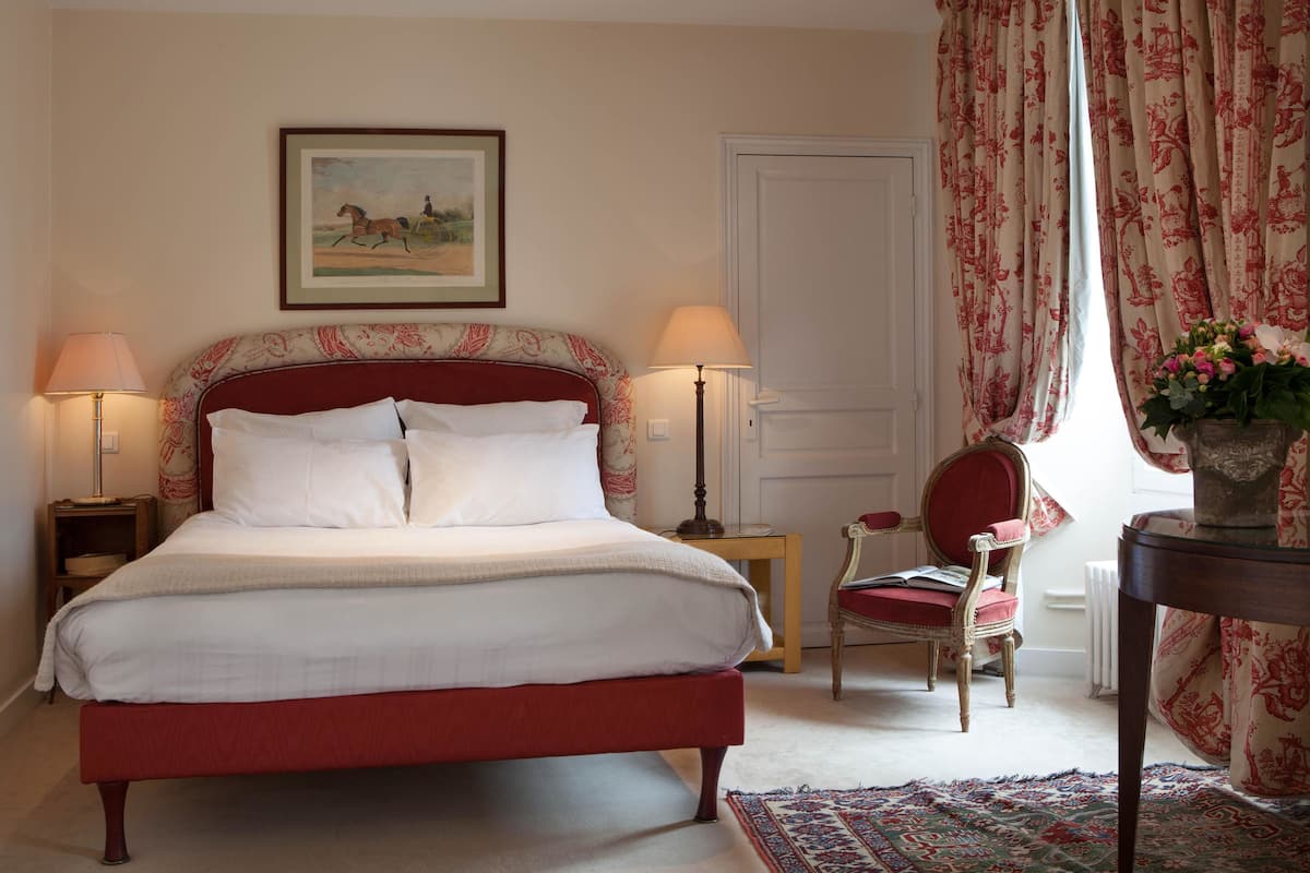 Een netjes opgemaakt bed met witte lakens, een rood hoofdeinde en twee bedlampjes. Daarboven hangt op de muur een ingelijst beeld, en er staat een rode stoel en een tafeltje met een bloevas.