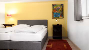 A bedroom with a double bed, a nightstand with a lamp, a small table, a colorful painting on the wall, and a red rug.