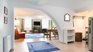 Un moderno salón con suelo de madera, un tapete azul, una zona de comedor con mesa y sillas, y una cocina con refrigerador y una estantería.