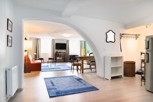 Ein modernes Wohnzimmer mit Holzboden, einem blauen Teppich, einem Essbereich mit Tisch und Stühlen sowie einer Küchenzeile mit Kühlschrank und einer Kleiderstange.