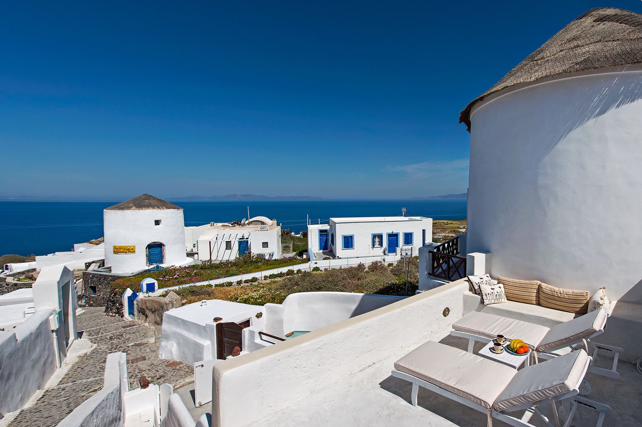 A white building with a blue door and a thatched roof, overlooking a coastal area with a clear blue sky.