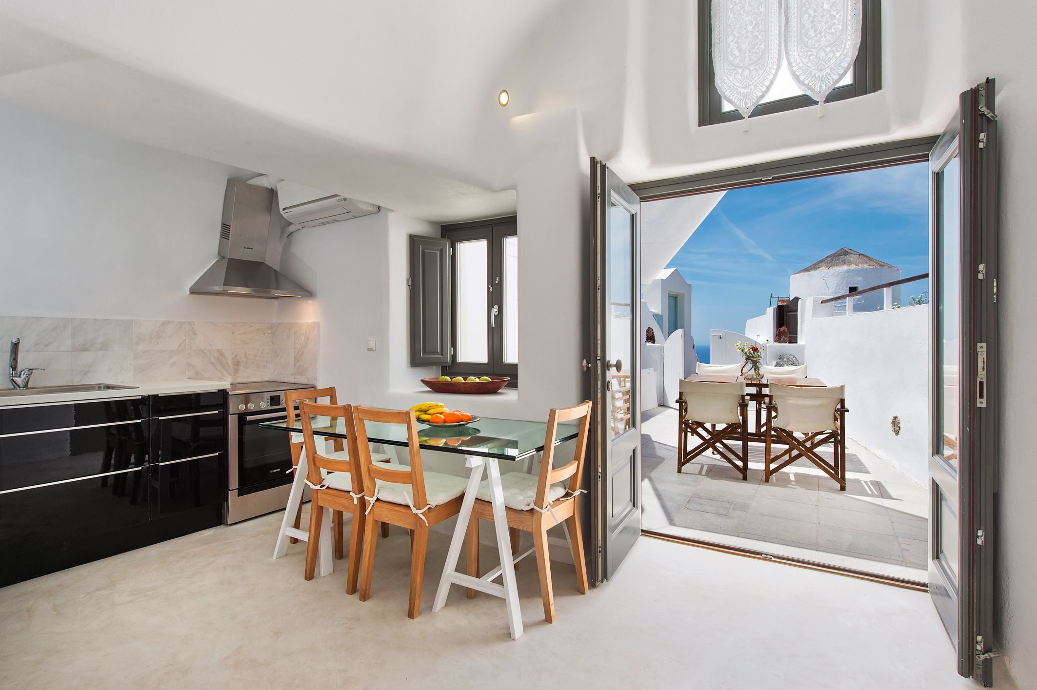 A modern kitchen with a glass dining table and chairs, a stainless steel range hood, and a view of the sea through an open door.