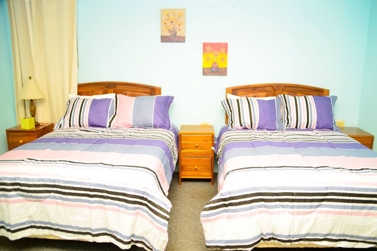 A twin room with two beds, each with striped bedding and pillows. There are wooden nightstands with lamps and a small drawer. The room has light blue walls and two framed pictures on the wall.