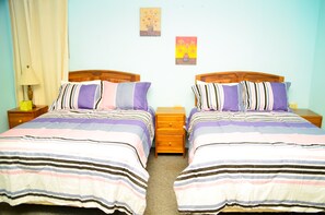 A twin room with two beds, each with striped bedding and pillows. There are wooden nightstands with lamps and a small drawer. The room has light blue walls and two framed pictures on the wall. - Wildgreen Residence (Point Fortin)