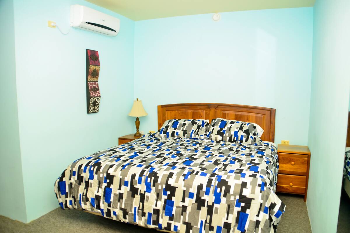 A bedroom with a bed, a wooden headboard, a nightstand, a lamp, and an air conditioning unit.