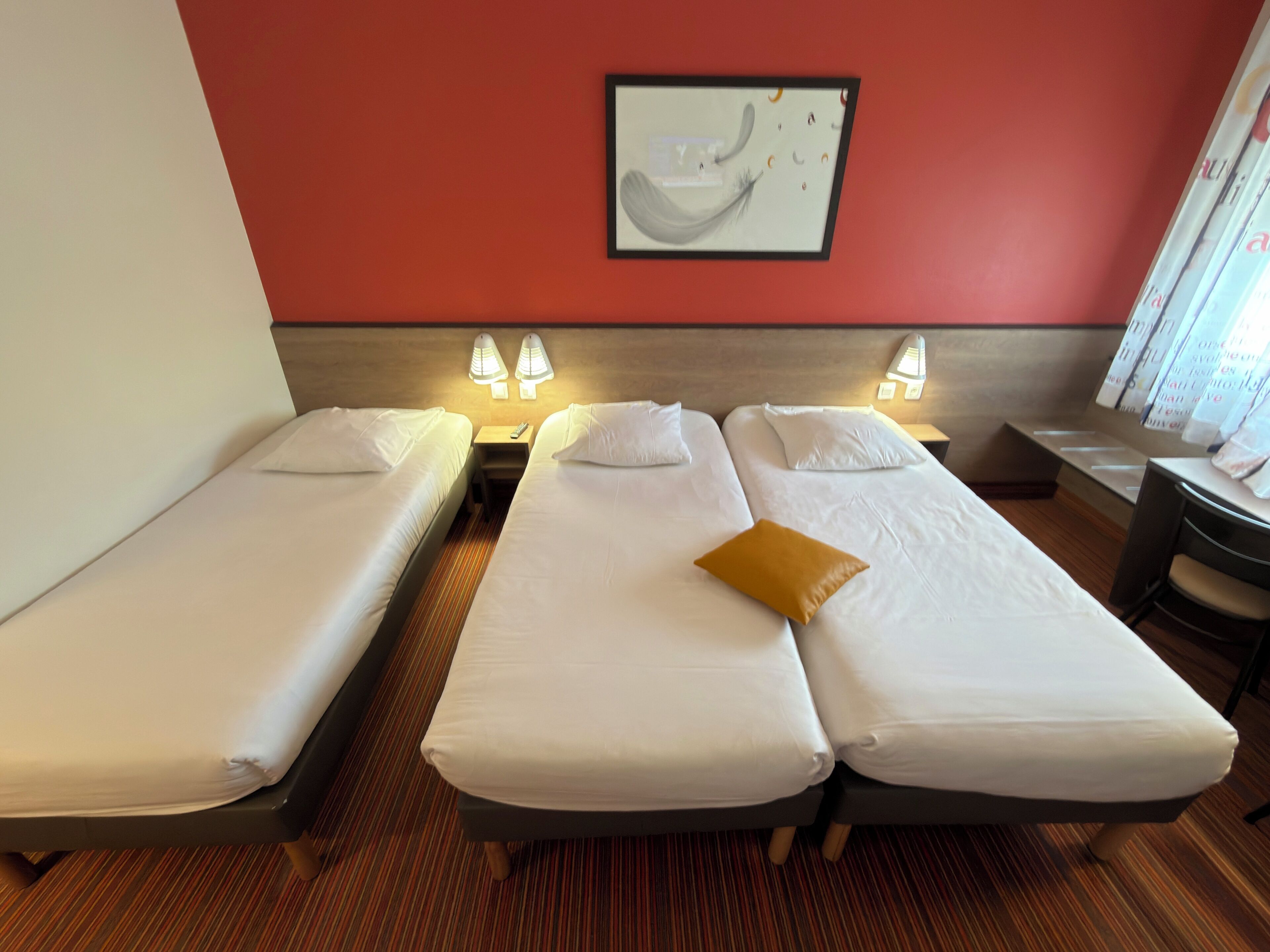 A hotel room with two single beds, a wooden floor, a red accent wall, and a framed abstract artwork.