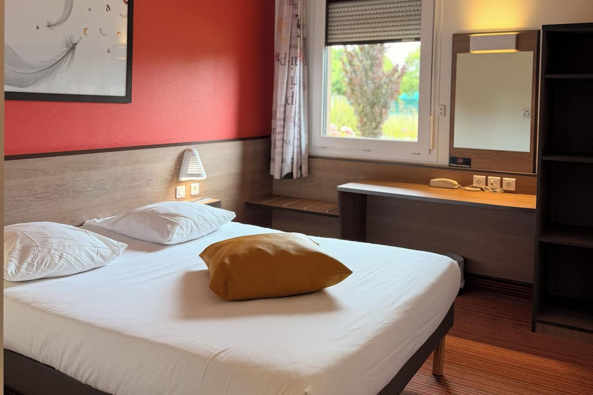 A hotel room with a bed, bedside table, lamp, window with blinds, and a mirror.