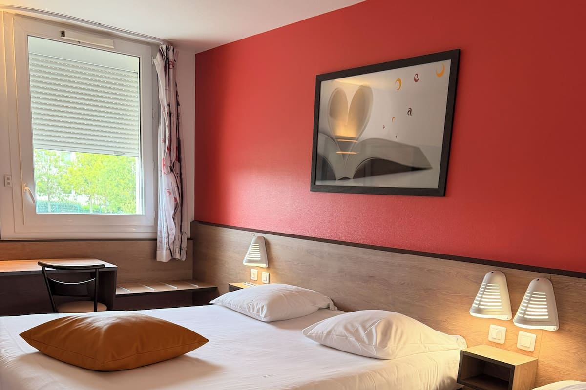 A hotel room with two beds, a desk, a chair, a window with curtains, and a wall-mounted picture.