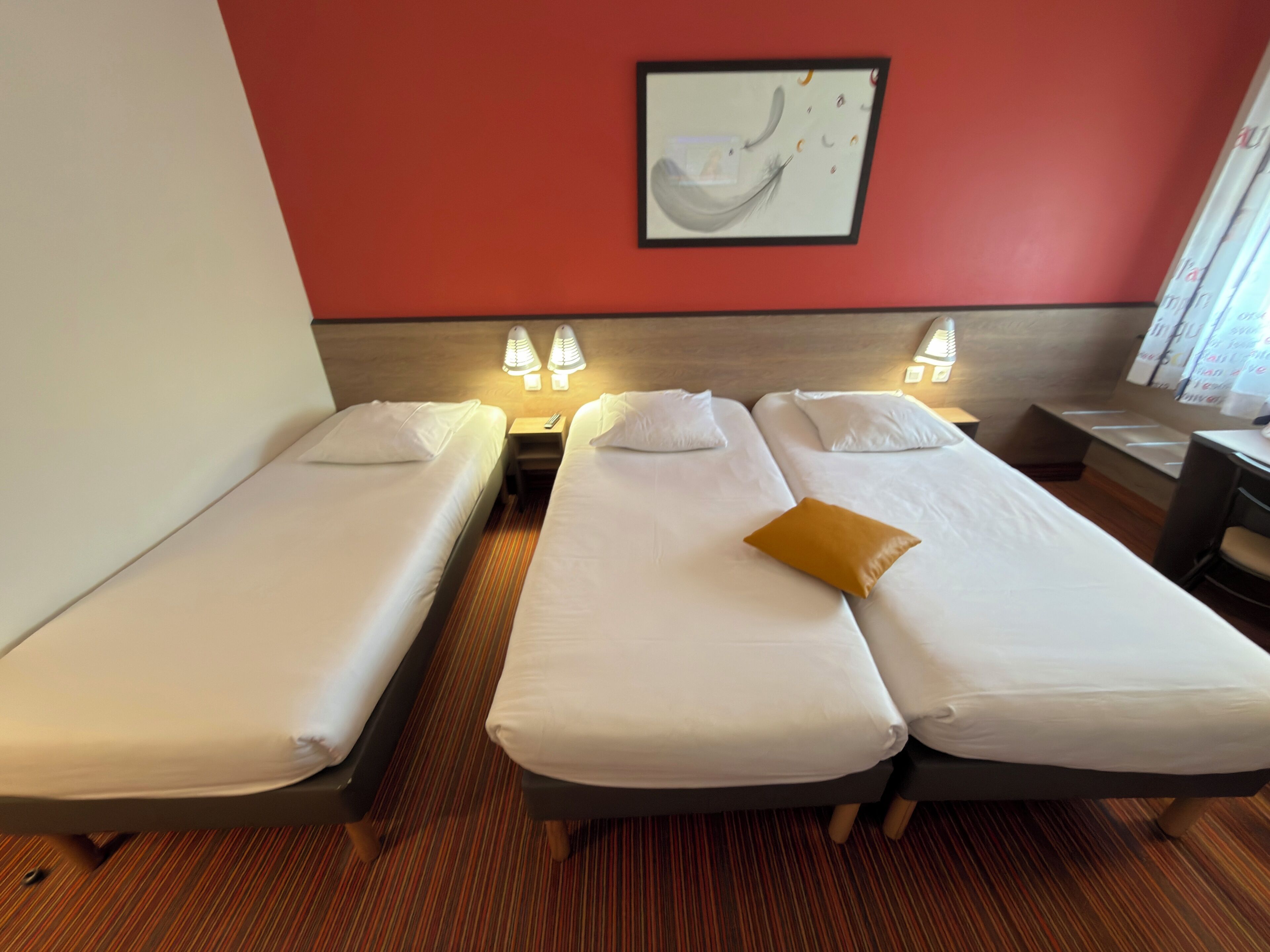 A hotel room with two single beds, a wooden headboard, a painting on the wall, and a desk with a chair.
