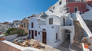 Un edificio tradicional de paredes blancas con una escalera que lleva a una puerta, rodeado de plantas en macetas y mobiliario de exterior.
