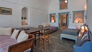 Una habitación acogedora y bien iluminada con una mesa de comedor de madera, un sofá y una cama. Hay dos ventanas con detalles azules, un estante con botellas y un cuadro en la pared.