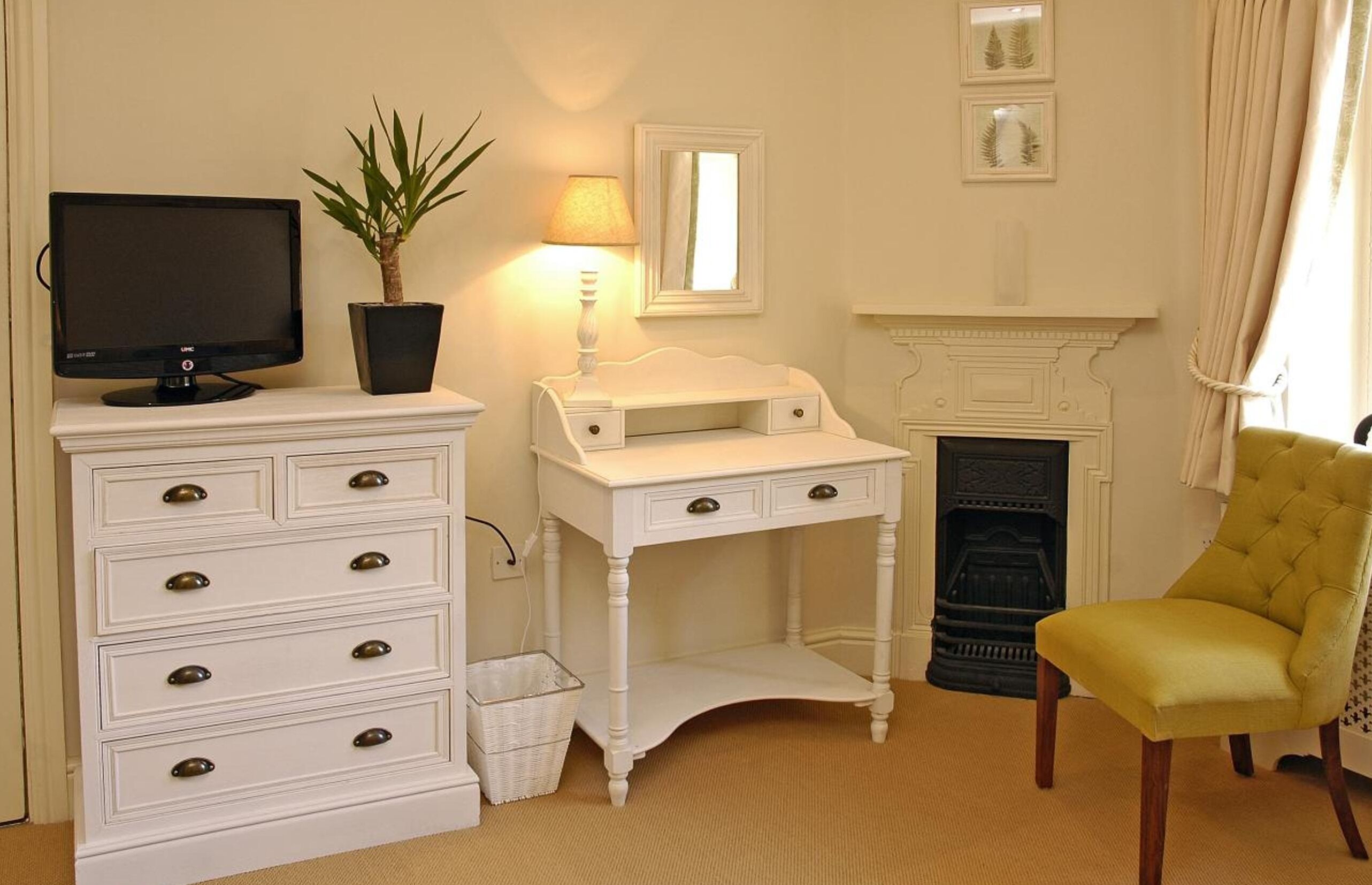 A room with a white dresser, a small white desk, a yellow armchair, a television, a potted plant, a fireplace, and framed pictures on the wall.