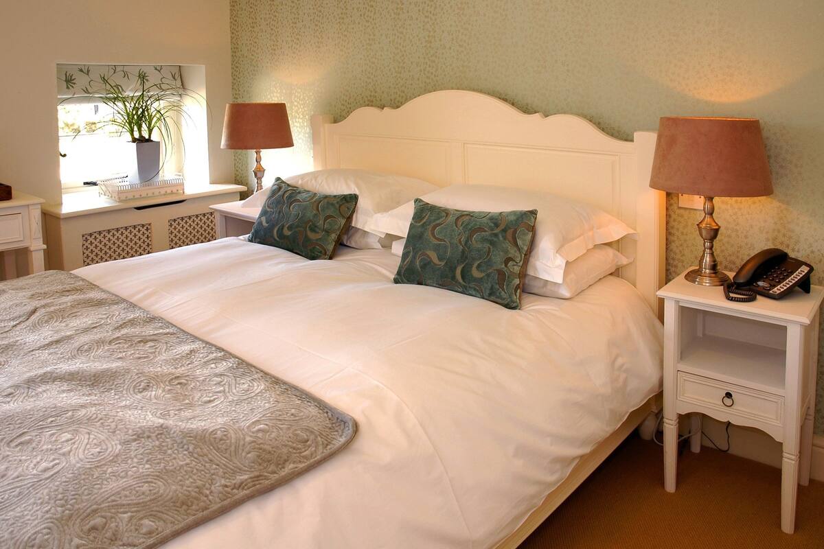 A neatly made bed with white linens and two decorative pillows, a bedside table with a lamp and a phone, and a window with a plant.