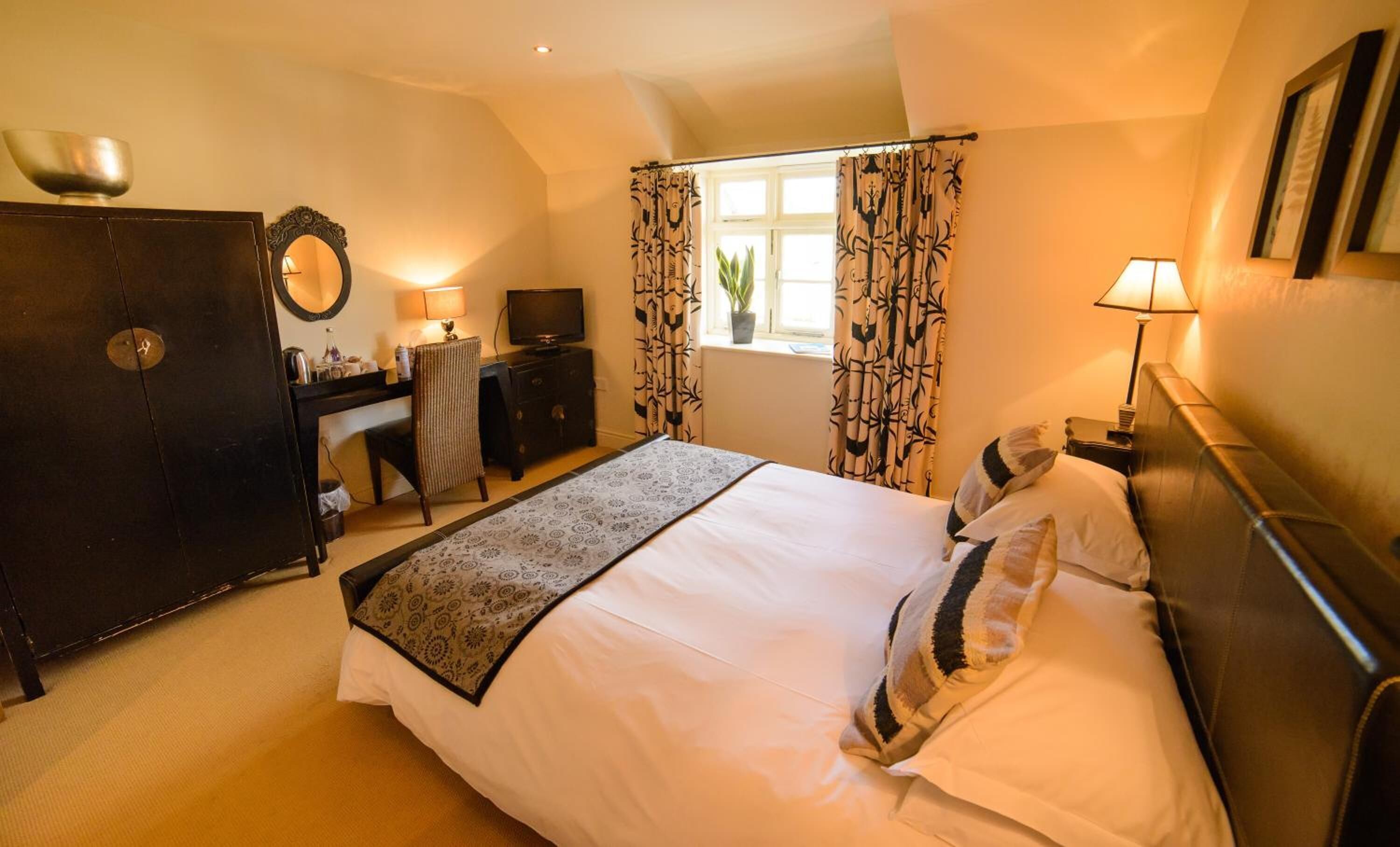 A hotel room with a bed, a desk, a chair, a TV, and a window with patterned curtains.