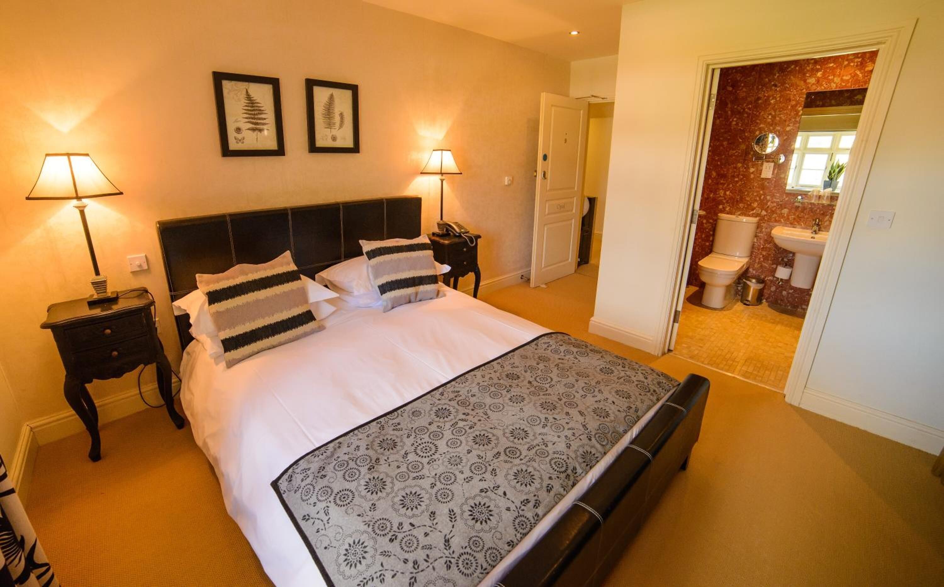 Habitación de hotel con una cama grande, dos mesitas de noche con lámparas, una puerta al baño e impresiones botánicas enmarcadas en la pared.