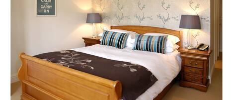 A neatly made bed with a brown headboard, two bedside tables with lamps, a wall with a floral wallpaper, and a sign that says "KEEP CALM AND CARRY ON".
