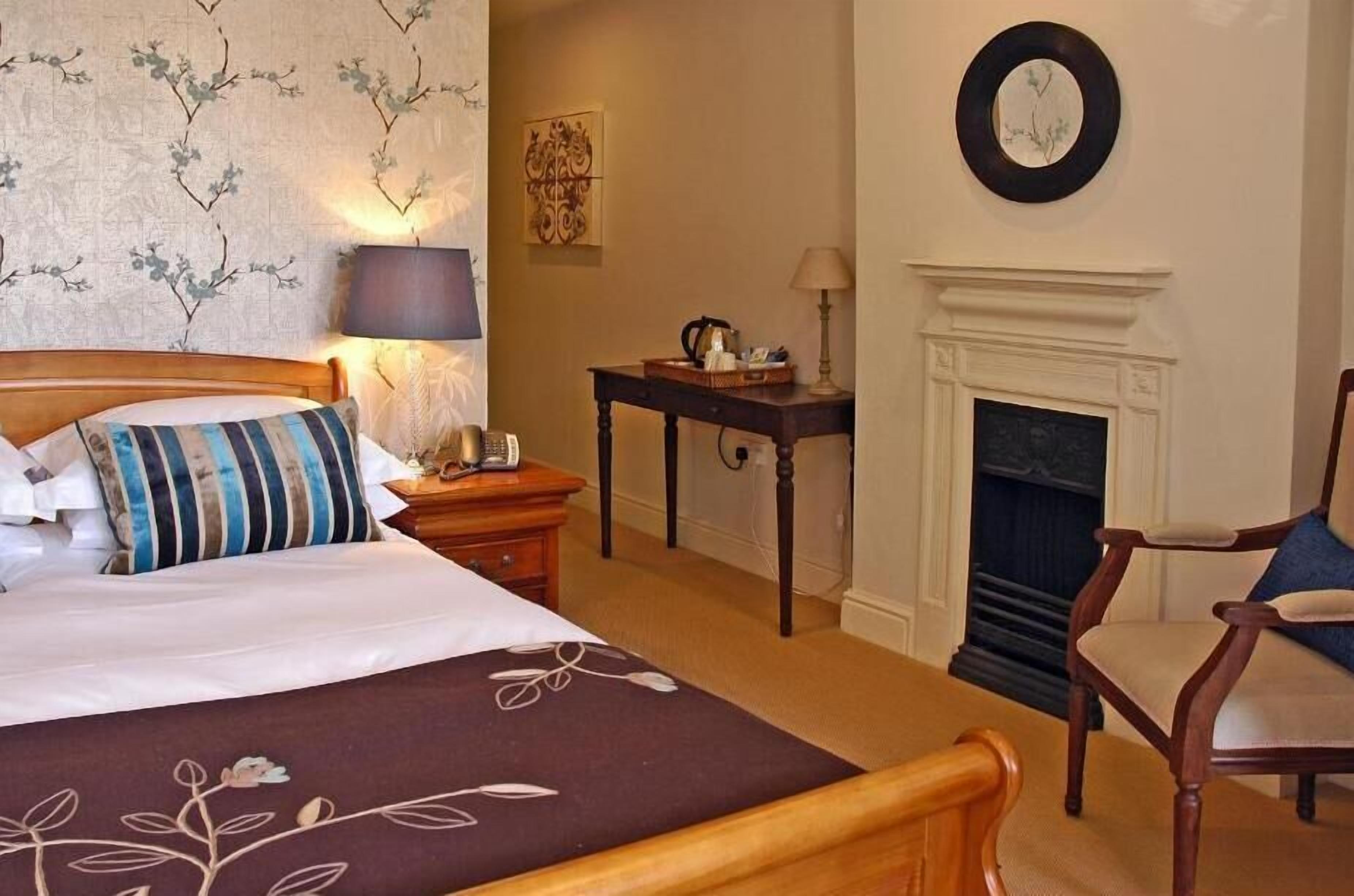 Una habitación de hotel con cama, una silla, una mesa con lámpara, chimenea y un reloj de pared.