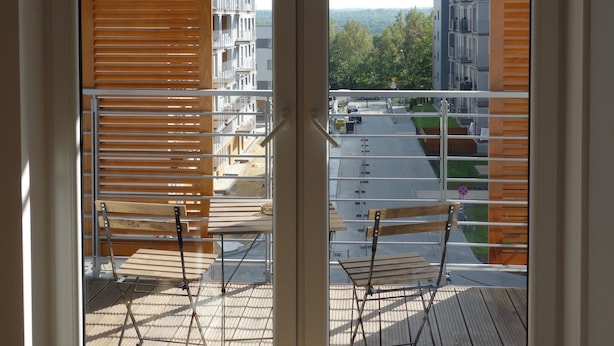 Näkymä ikkunasta, jossa on puusellaiset sälekaihtimet. Ikkunasta avautuu näkymä parvekkeelle, jossa on tuoleja ja pöytä, ja taustalla näkyy rakennuksia.