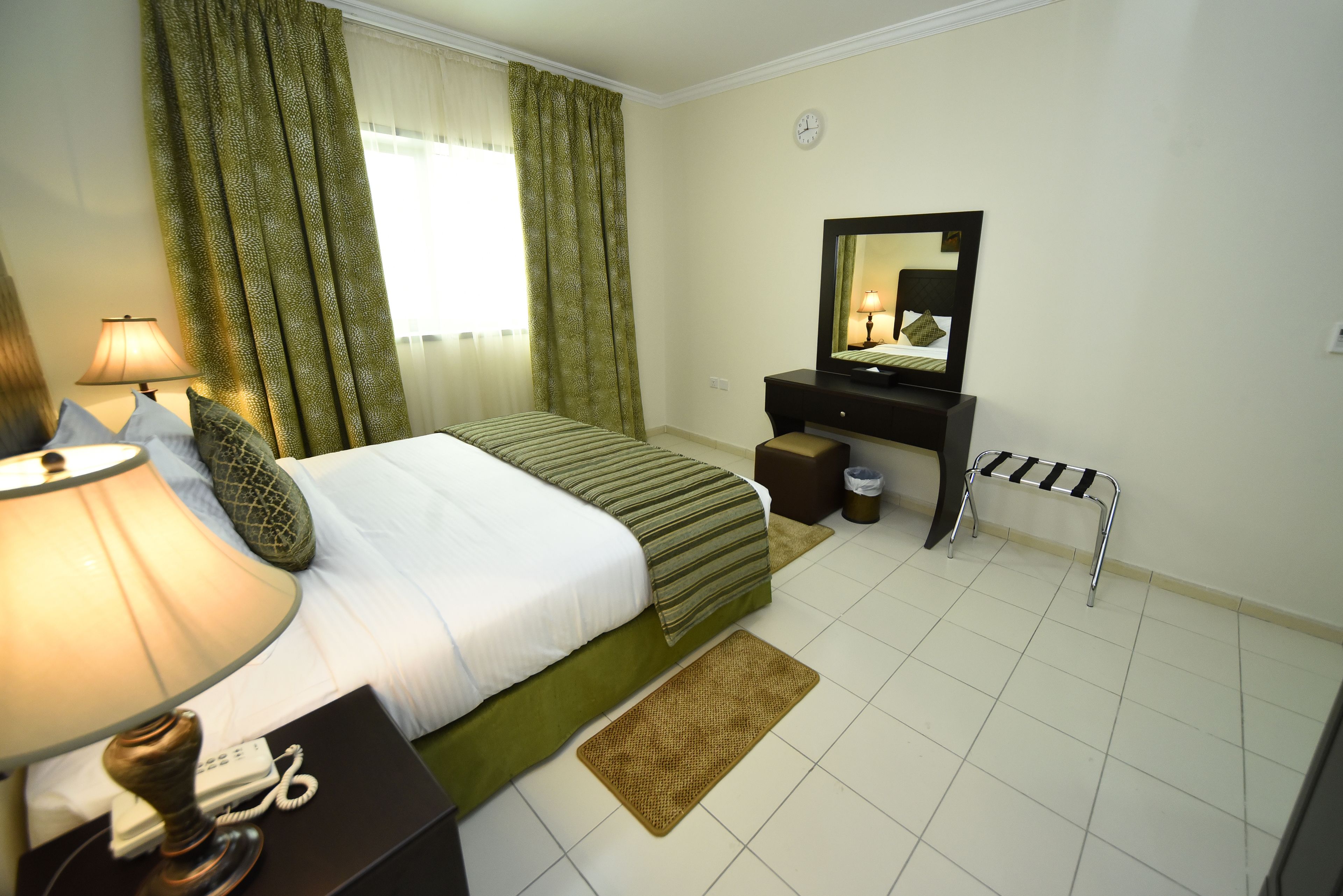 A hotel room with a bed, bedside lamp, a small table with a lamp, a mirror, a trash bin, and a small stool.