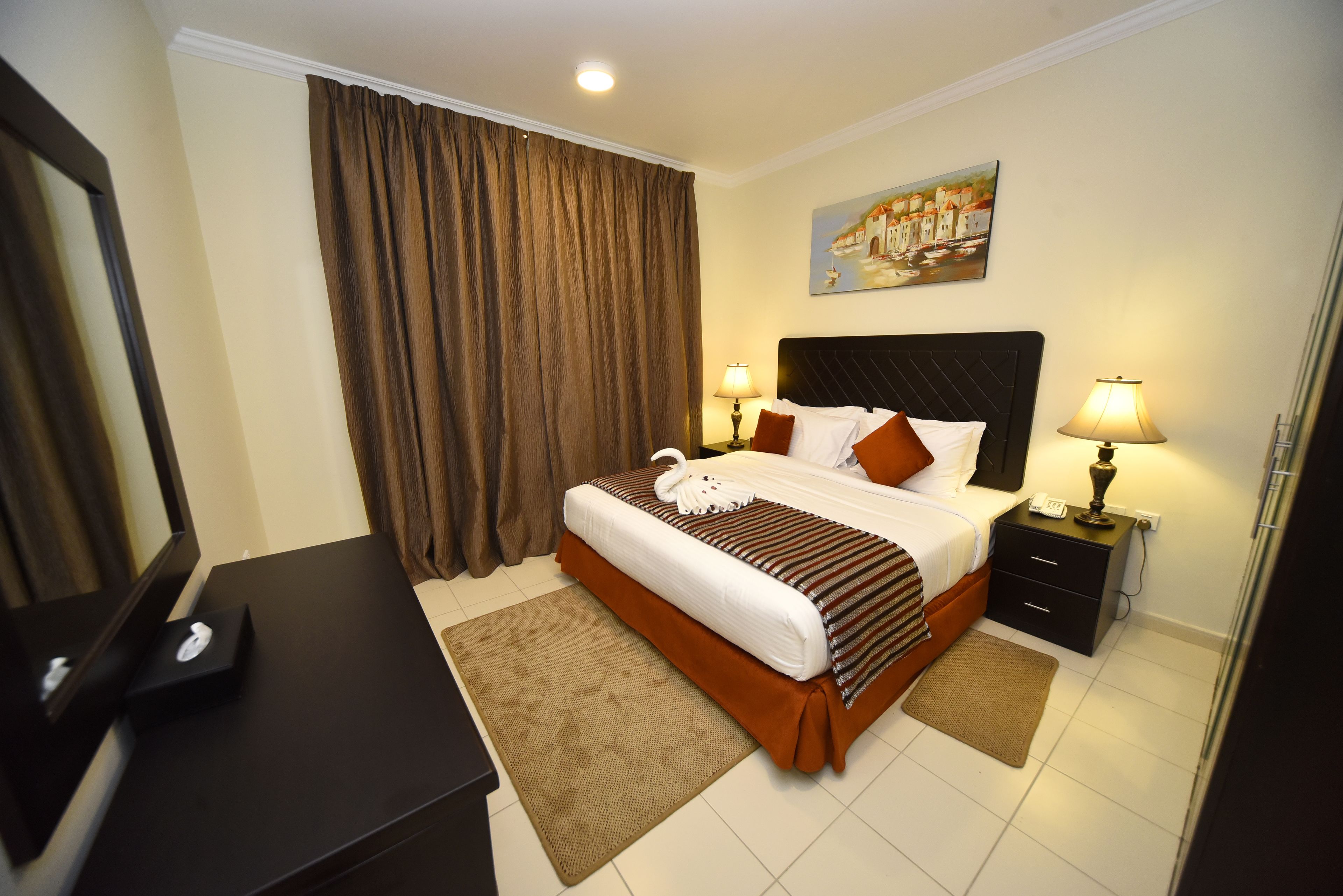 A hotel room with a bed, two bedside tables with lamps, a painting on the wall, and a desk with a telephone.