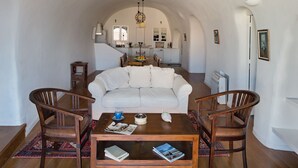 A cozy living room with a white sofa, wooden coffee table, and two wooden chairs. A dining table with chairs is in the background. - Villa Amalia (Santorini)