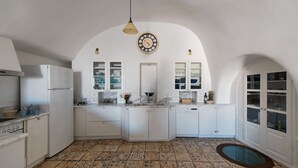 A kitchen with white cabinets, a white refrigerator, a clock, and a tiled floor. - Villa Amalia (Santorini)
