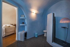 A bathroom with a blue wall, a shower, a toilet, and a bed in the adjacent room. - Villa Amalia (Santorini)