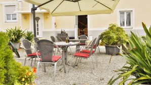 Een terras buiten met een tafel, stoelen, een grote parasol en potplanten.