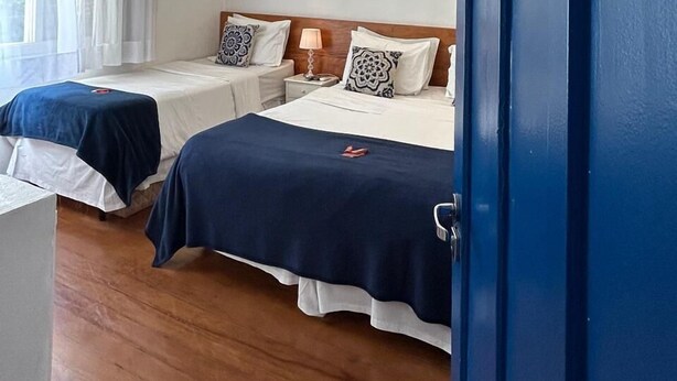 Een hotelkamer met twee bedden, een houten vloer, een raam met witte gordijnen en een blauwe deur.