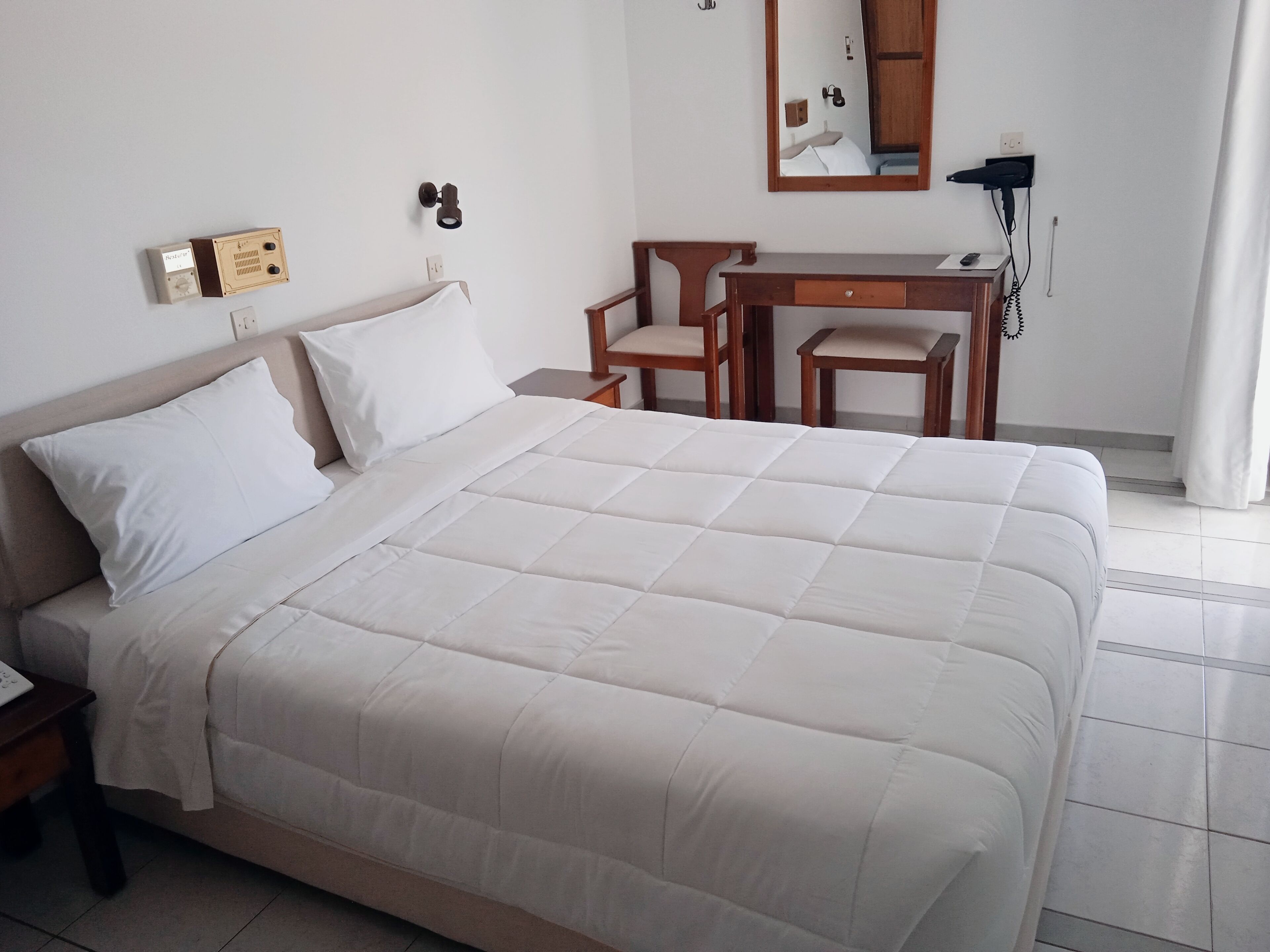 A neatly made bed with white bedding, a wooden nightstand, a desk with a chair, and a telephone.