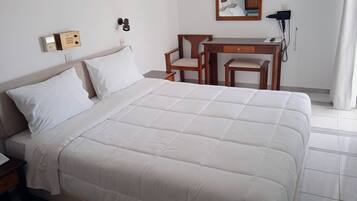A neatly made bed with white bedding, a wooden nightstand, a desk with a chair, and a telephone.