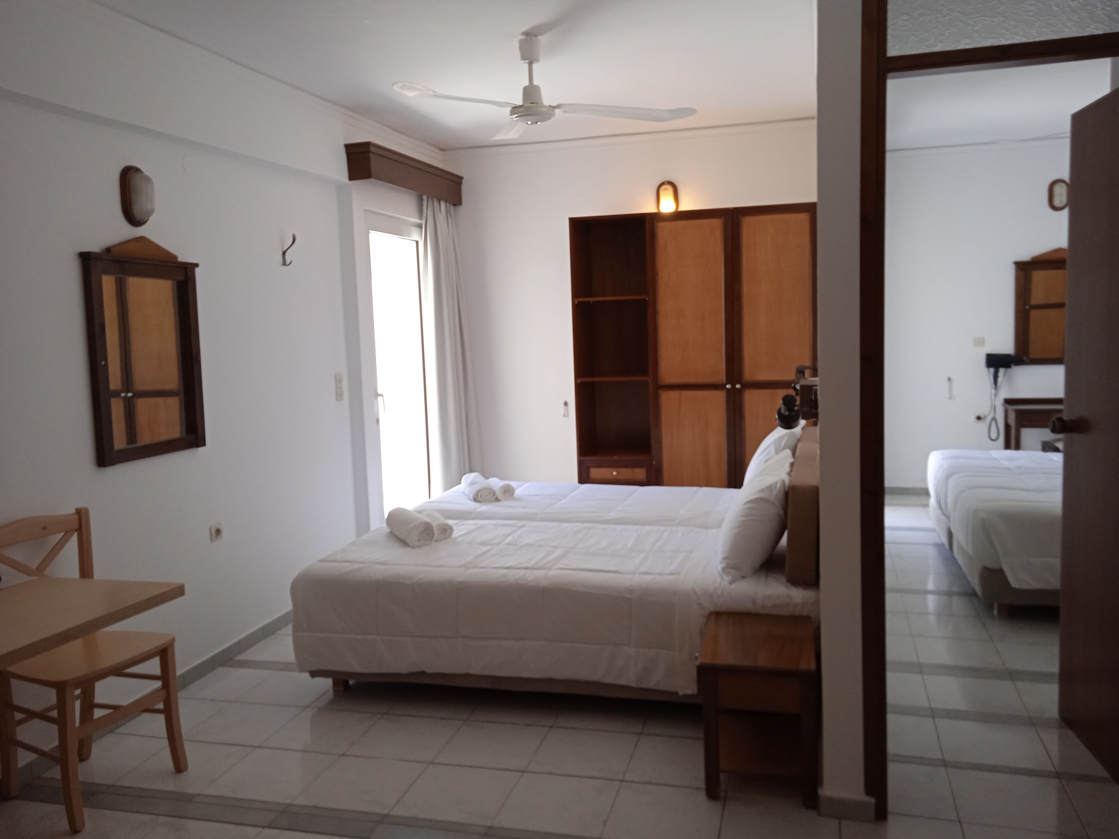 A bedroom with a bed, a desk, and a chair. There is a wardrobe and a ceiling fan.