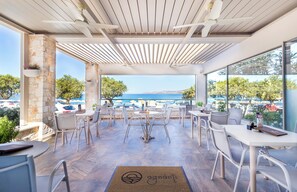 An outdoor seating area with tables and chairs, a view of the sea, and a stone pillar. - Pal Beach (Kantanos-Selino)