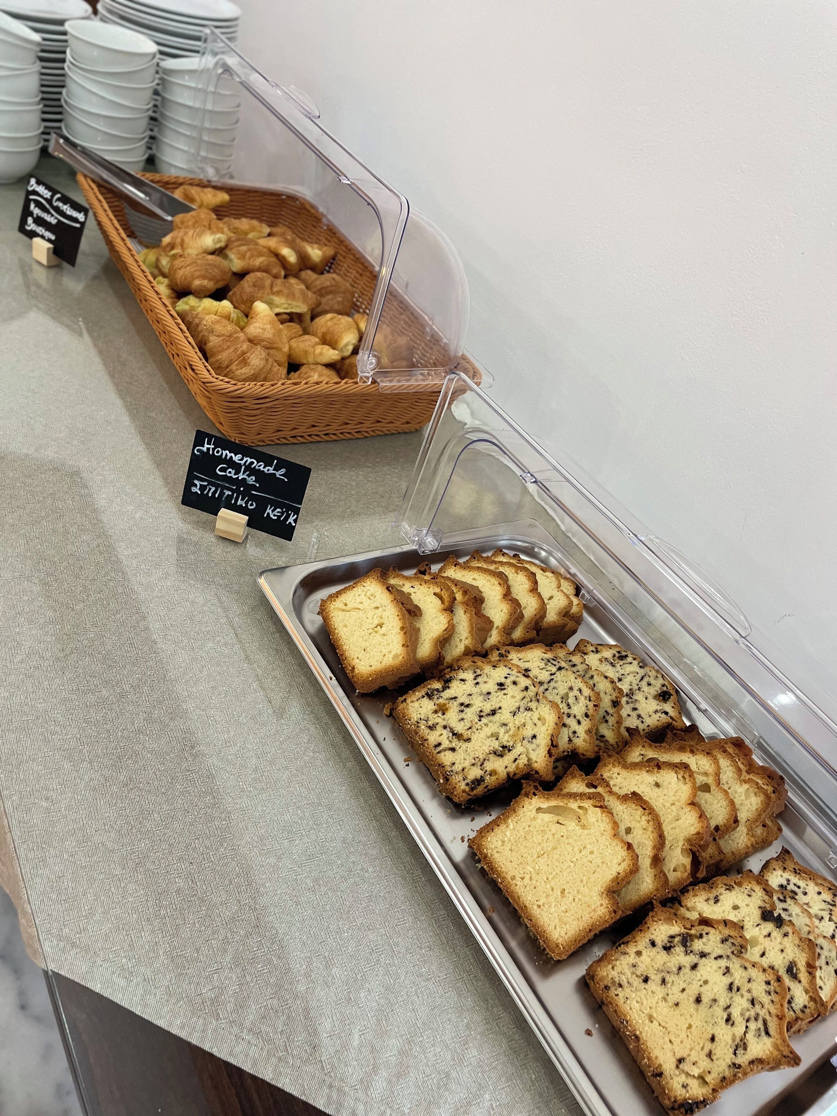 Un mostrador de buffet con bandejas de pan y bollería, incluyendo croissants y un cartel que indica tarta casera.