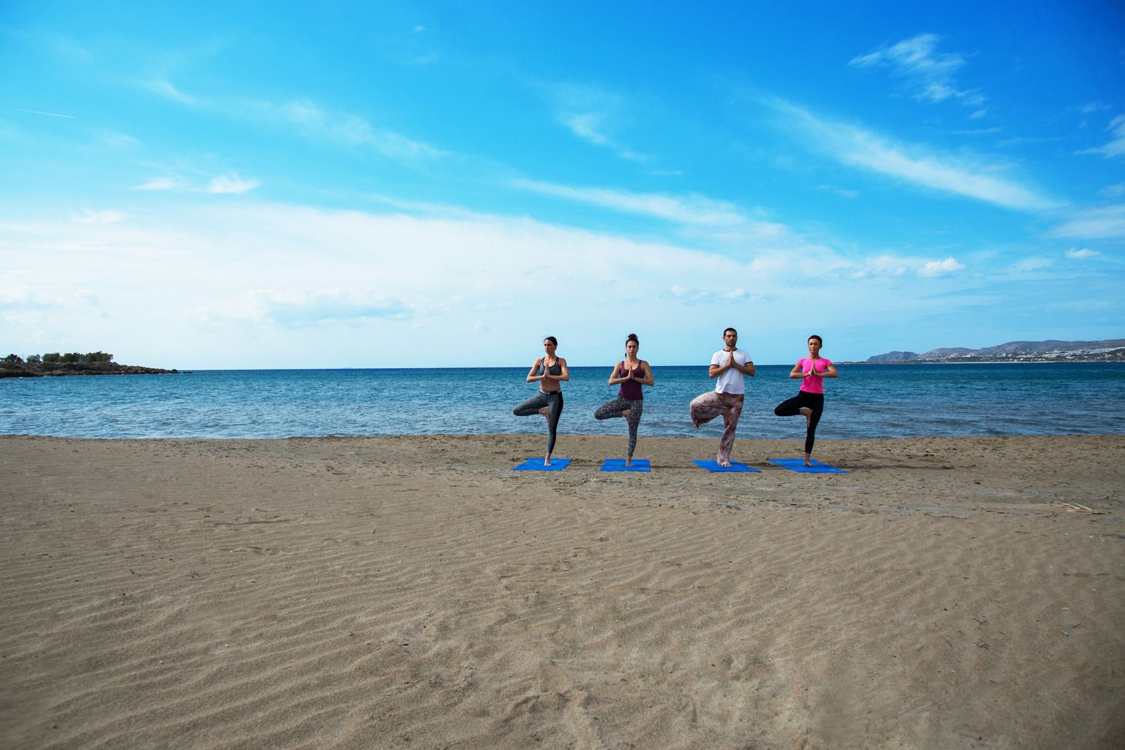Four individuals practicing yoga on blue mats on a sandy beach with a calm sea and clear sky in the background.
