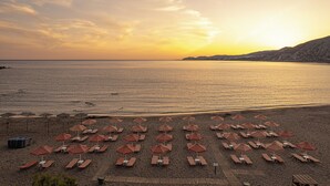 Una playa con hileras de tumbonas y sombrillas, una terraza de madera y vistas al mar y colinas lejanas.